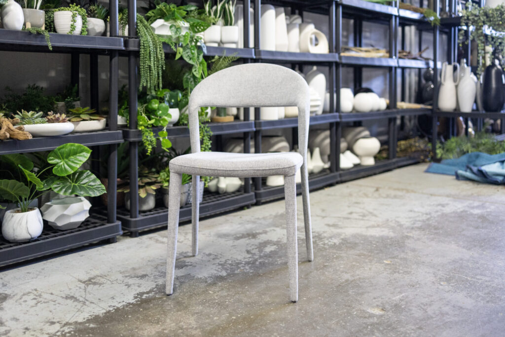 Unwrapped chair in a storage area with plants and decor items on shelves in the background.