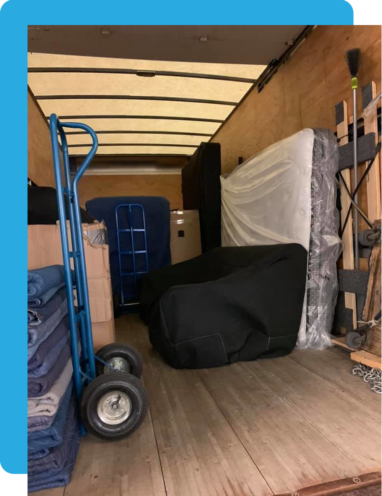 Inside view of a moving truck prepared by Toronto movers, showing items packed for transportation. A dolly with large wheels is positioned on the left, next to stacks of folded moving blankets. Several large items, including a mattress and an armchair covered in protective wrapping, are secured within the truck. Additional packing materials and tools are visible on the sides, with a broom stored upright on the right.