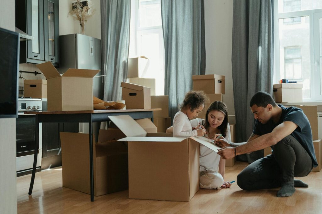 A family of three packing and labeling cardboard boxes in a modern Toronto apartment, preparing for their move. The room is filled with moving boxes, and the parents are helping their child decorate a box.