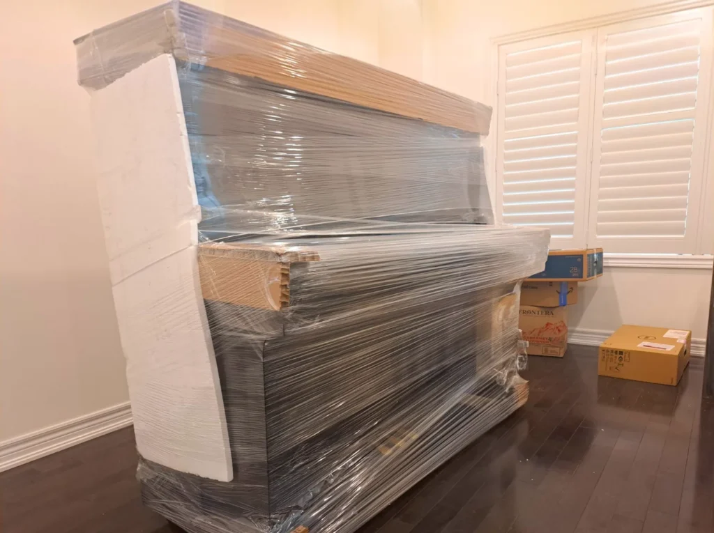 多伦多安达搬运为黑色立式钢琴包裹高密度泡沫板和透明薄膜，提供全方位缓冲保护，避免运输中震动损伤。